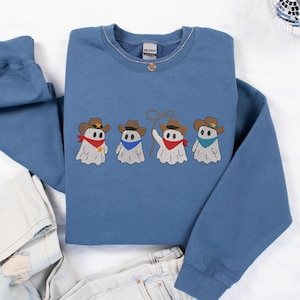 May include: A blue crewneck sweatshirt featuring an embroidered graphic of four white ghosts wearing brown cowboy hats. The ghosts are adorned with red or blue bandanas, and one ghost holds a lasso. The sweatshirt has long sleeves and a ribbed collar.
