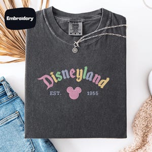 a black t-shirt with a Disneyland logo, a pair of blue jeans, and some accessories such as a necklace and a scarf, all laid out on a white surface.