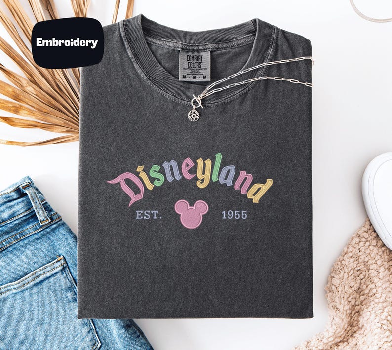 a black t-shirt with a Disneyland logo, a pair of blue jeans, and some accessories such as a necklace and a scarf, all laid out on a white surface.
