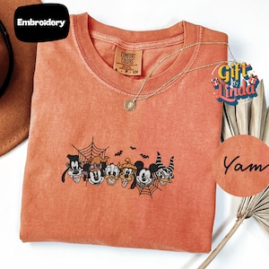 May include: A burnt orange t-shirt featuring an embroidered design of various cartoon characters wearing Halloween costumes, including witch hats and horns, accented by spider webs and small bats. The shirt has a crew neckline and a Comfort Colors label.