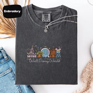 a gray t-shirt with a graphic design of Walt Disney World, including various Disney landmarks and attractions. The t-shirt is laid out on a white surface, along with a pair of jeans and a necklace.