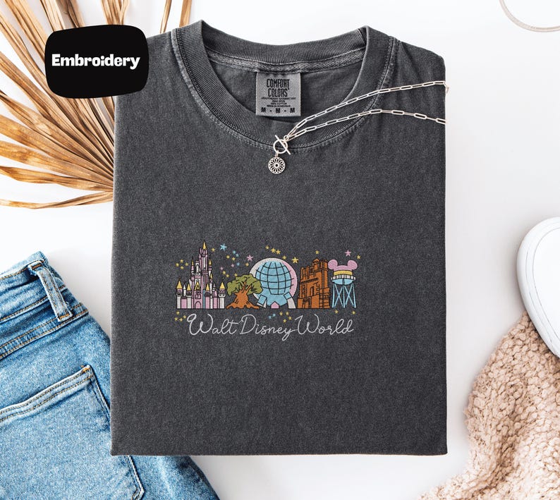 a gray t-shirt with a graphic design of Walt Disney World, including various Disney landmarks and attractions. The t-shirt is laid out on a white surface, along with a pair of jeans and a necklace.