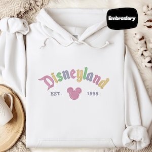 A white hoodie with a Disneyland logo and text, along with a mug and some cotton balls, is displayed on a surface.