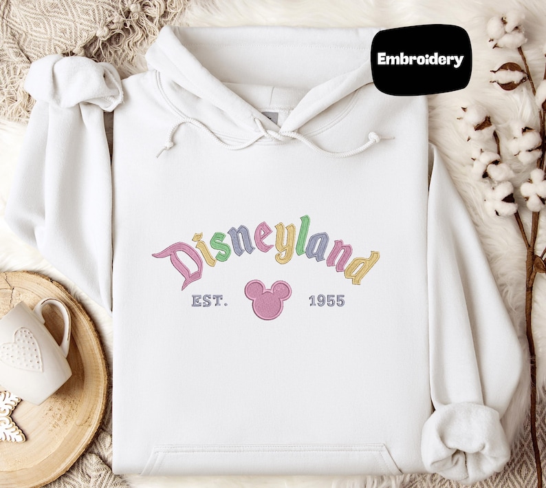 A white hoodie with a Disneyland logo and text, along with a mug and some cotton balls, is displayed on a surface.