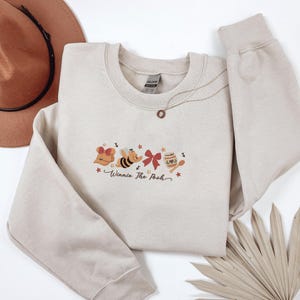 May include: A beige sweatshirt featuring an embroidered design of Winnie the Pooh characters, including Pooh Bear, a bee, a red bow, and a honey pot. The words "Winnie The Pooh" are written in a script font below the characters. A brown hat and necklace are also visible.