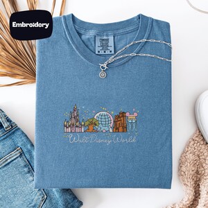 a blue t-shirt with a Disney World-themed embroidered design, along with a pair of jeans and a necklace.