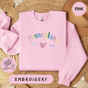 A pink sweatshirt with the word "Disneyland" embroidered on it, along with a straw hat and a straw hat. The sweatshirt is displayed on a woven mat, and there is a label that says "EMBROYERY" below the sweatshirt.