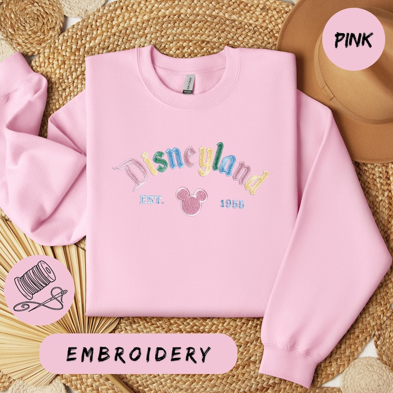 A pink sweatshirt with the word "Disneyland" embroidered on it, along with a straw hat and a straw hat. The sweatshirt is displayed on a woven mat, and there is a label that says "EMBROYERY" below the sweatshirt.