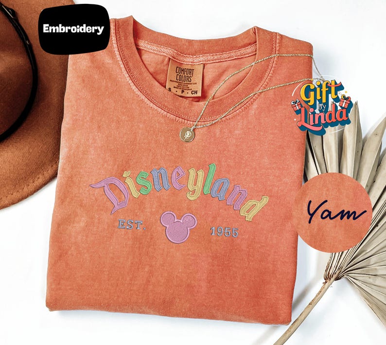 a pink t-shirt with the text "Disneyland EST. 1955" and a Mickey Mouse logo, along with a necklace and a palm frond.
