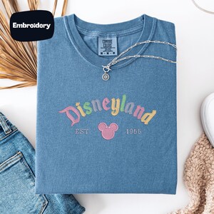 a blue t-shirt with the text "Disneyland EST. 1955" embroidered on it, along with a Mickey Mouse logo. The t-shirt is laid out on a white surface, accompanied by a pair of jeans, a necklace, and a sweater.