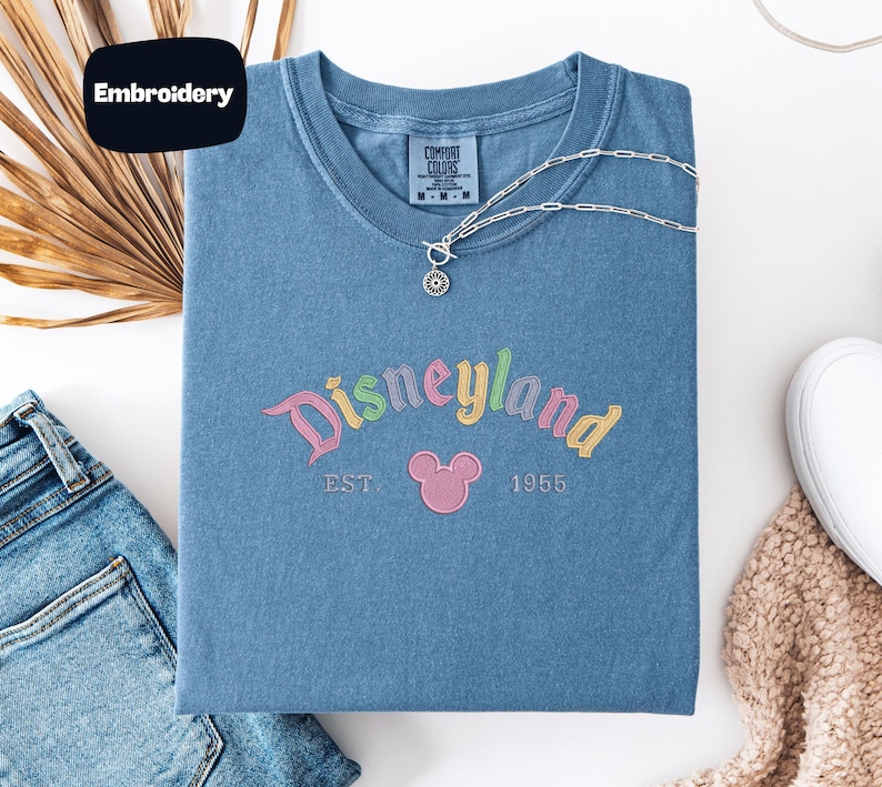 a blue t-shirt with the text "Disneyland EST. 1955" embroidered on it, along with a Mickey Mouse logo. The t-shirt is laid out on a white surface, accompanied by a pair of jeans, a necklace, and a sweater.