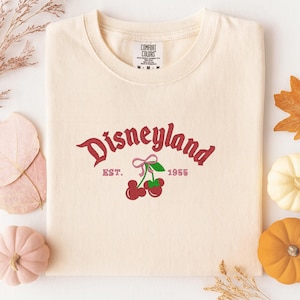 A white t-shirt with a Disneyland logo and text, along with autumn-themed decorations such as leaves, pumpkins, and gourds.