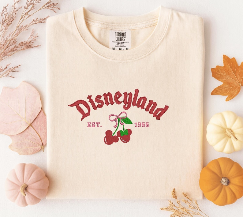 A white t-shirt with a Disneyland logo and text, along with autumn-themed decorations such as leaves, pumpkins, and gourds.