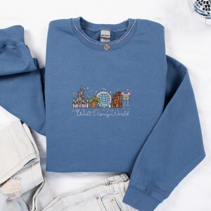 A blue sweatshirt with a graphic design of various Disney landmarks and characters, including the Eiffel Tower, Walt Disney World, and Mickey Mouse, is displayed on a white background.