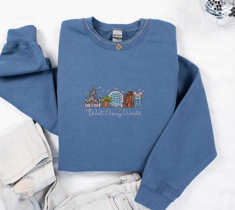 A blue sweatshirt with a graphic design of various Disney landmarks and characters, including the Eiffel Tower, Walt Disney World, and Mickey Mouse, is displayed on a white background.