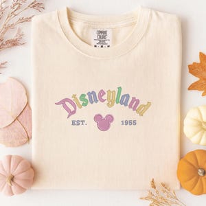 a white t-shirt with a Disneyland logo, surrounded by autumn-themed decorations such as leaves, pumpkins, and gourds.