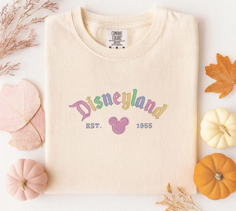 a white t-shirt with a Disneyland logo, surrounded by autumn-themed decorations such as leaves, pumpkins, and gourds.