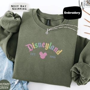 A green sweatshirt with the text "Disneyland EST. 1955" printed on it, along with a small Mickey Mouse logo. The sweatshirt is displayed on a white background, and there is a "Next Day Shipping" label and a "Military Green" label visible.