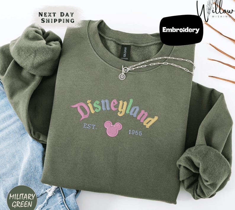 A green sweatshirt with the text "Disneyland EST. 1955" printed on it, along with a small Mickey Mouse logo. The sweatshirt is displayed on a white background, and there is a "Next Day Shipping" label and a "Military Green" label visible.