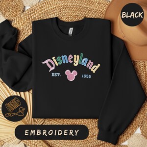 A black sweatshirt with the text "Disneyland EST. 1955" embroidered on it, along with a Mickey Mouse logo. The sweatshirt is laid out on a woven straw mat, and there is a hat and a straw hat visible in the background.