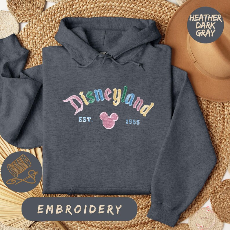 A gray hoodie with the word "Disneyland" embroidered on it, along with a hat and straw hat, all laid out on a woven mat.