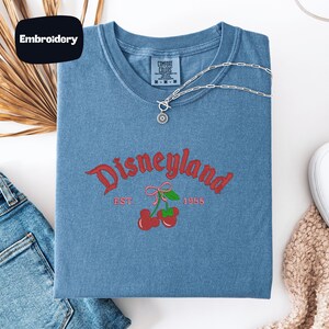 a blue t-shirt with a Disneyland logo, a denim shirt, a pair of jeans, and a beige sweater, all laid out on a white surface.