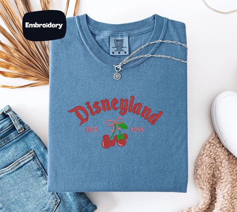a blue t-shirt with a Disneyland logo, a denim shirt, a pair of jeans, and a beige sweater, all laid out on a white surface.
