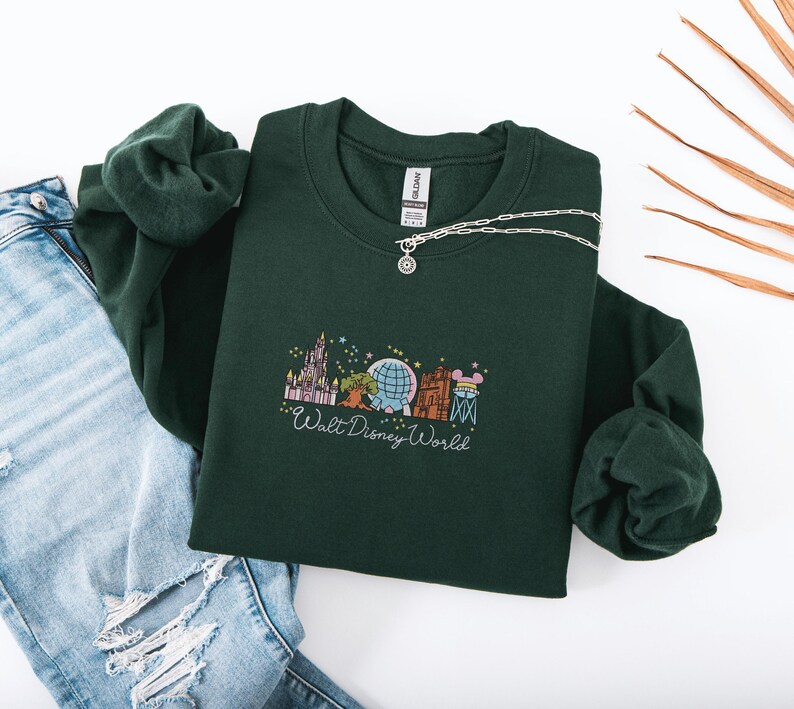 a green sweater with a Disney World logo, a necklace, and a pair of blue jeans, all laid out on a white background.