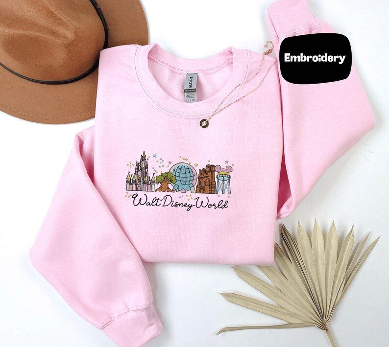 A pink sweatshirt with a Disney World logo, a brown hat, and some leaves are arranged on a white background.