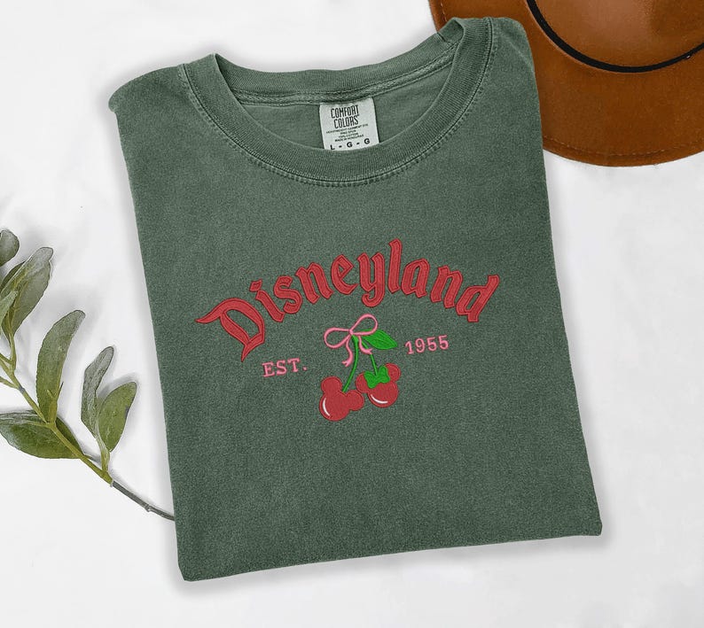 A green t-shirt with the text "Disneyland EST. 1955" printed on it, along with a red bow and cherries, is displayed on a white surface.