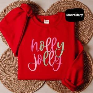 May include: A bright red long-sleeve crewneck sweatshirt featuring the text "holly jolly" embroidered in a cursive font. The letters alternate between shades of pink and light green. A black tag in the upper right corner displays the word "Embroidery".