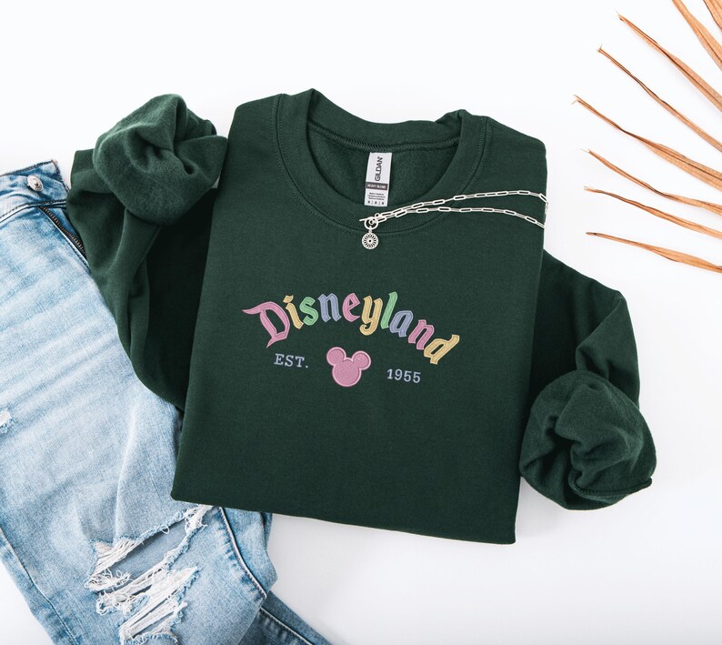 A green sweatshirt with the text "Disneyland EST. 1965" printed on it, along with a small Mickey Mouse logo, is displayed on a white background.