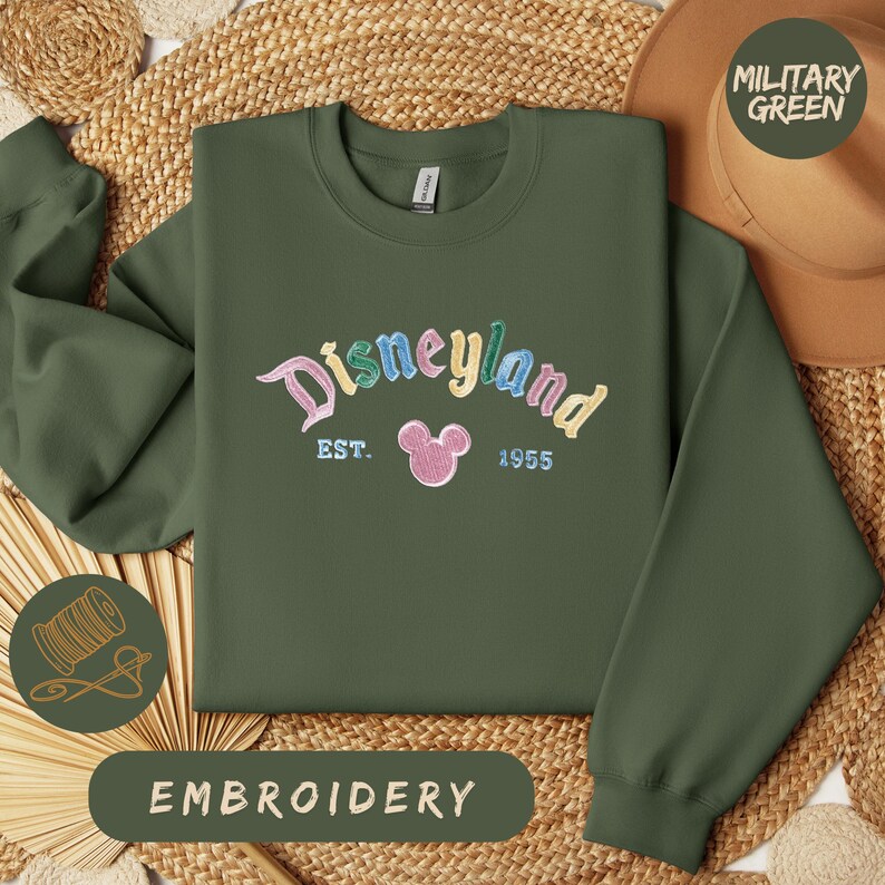 a green sweatshirt with the word "Disneyland" embroidered on it, along with the text "EST. 1955" and a pink Mickey Mouse logo. The sweatshirt is laid out on a woven mat, and there is a brown hat and a straw hat in the background. The image also includes the text "EMBROIDERY" and "MILITARY GREEN".