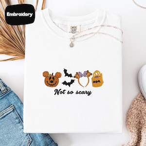 a white t-shirt with a Halloween-themed embroidered design, including a pumpkin, bats, and a bag, along with the text "Not so scary".