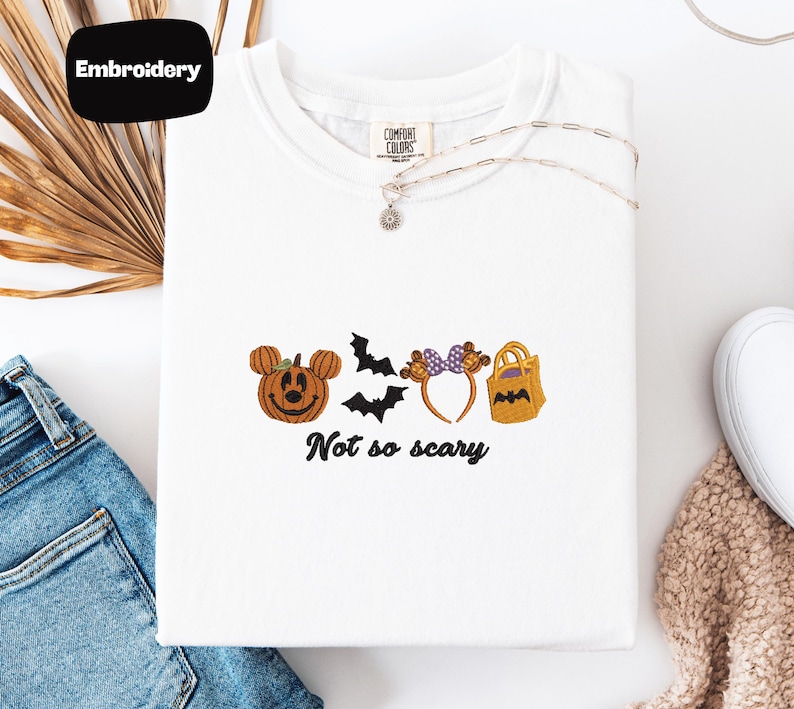 a white t-shirt with a Halloween-themed embroidered design, including a pumpkin, bats, and a bag, along with the text "Not so scary".