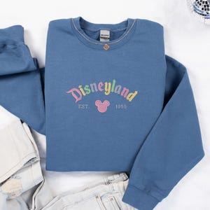 A blue sweatshirt with the text "Disneyland EST. 1955" printed on it, along with a Mickey Mouse logo, is laid out on a white surface.