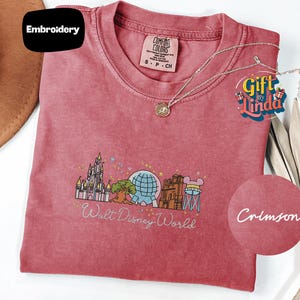 a pink t-shirt with a Disney World-themed design, including a castle, a globe, and various Disney characters. The t-shirt is accompanied by a necklace and a gift tag, suggesting it is a merchandise item related to Disney.