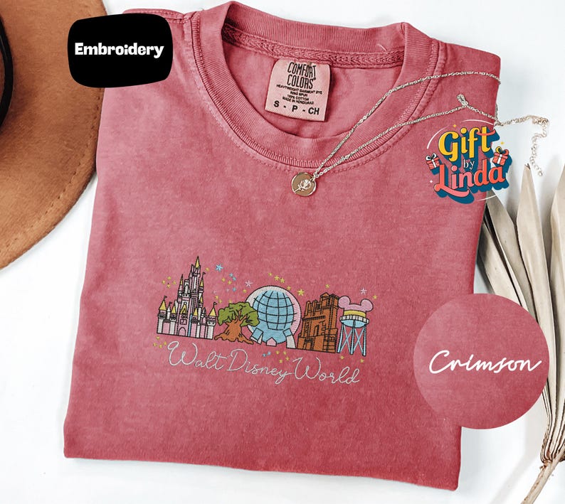 a pink t-shirt with a Disney World-themed design, including a castle, a globe, and various Disney characters. The t-shirt is accompanied by a necklace and a gift tag, suggesting it is a merchandise item related to Disney.