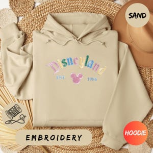 A beige hoodie with the word "Disneyland" embroidered on it, along with a hat and straw hat, all laid out on a woven surface.