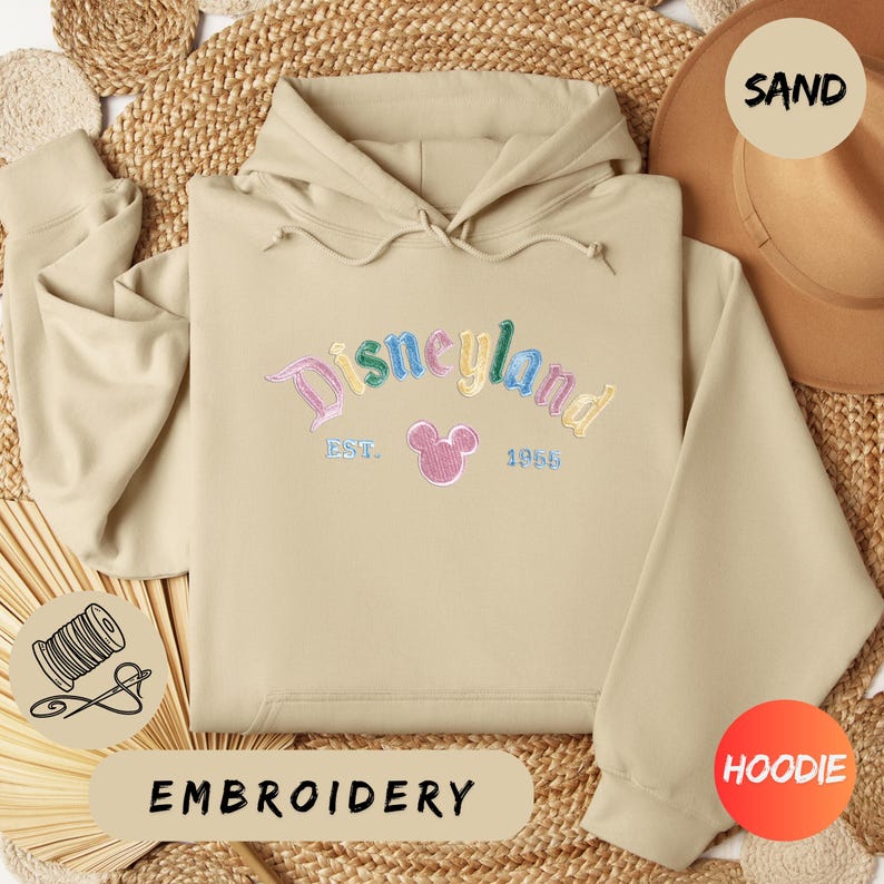 A beige hoodie with the word "Disneyland" embroidered on it, along with a hat and straw hat, all laid out on a woven surface.