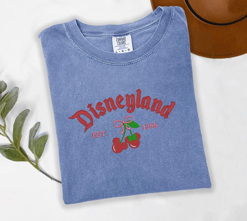 A blue t-shirt with the text "Disneyland EST. 1955" printed on it, along with a red cherry graphic, is displayed on a white surface.