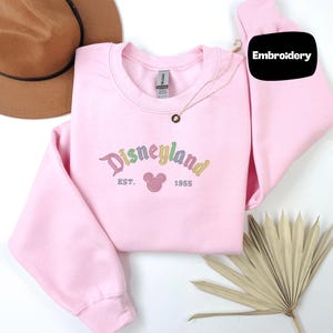 a pink sweater with the text "Disneyland EST. 1955" embroidered on it, along with a brown hat and a fan.