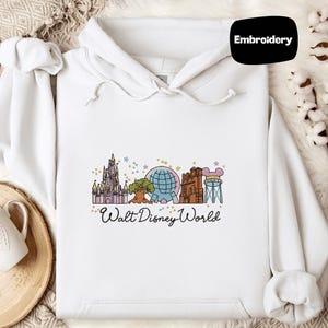 A white hoodie with a Disney World-themed embroidered design, including various landmarks and attractions, is displayed alongside a mug and a small plant.