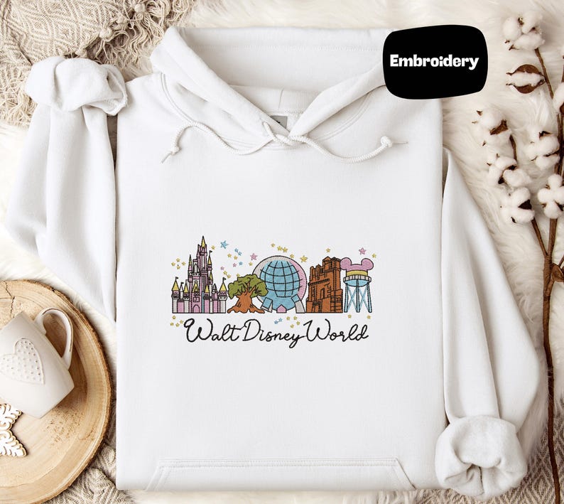 A white hoodie with a Disney World-themed embroidered design, including various landmarks and attractions, is displayed alongside a mug and a small plant.