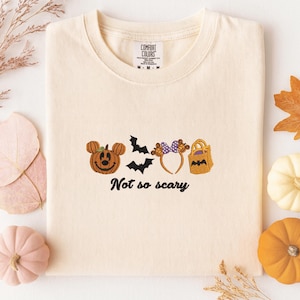 A white t-shirt with a Halloween-themed design, including a pumpkin, bats, and a bag, along with the text "Not so scary" printed on it. The t-shirt is surrounded by autumn decorations such as leaves and pumpkins.
