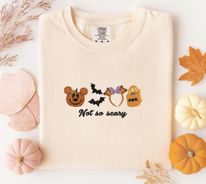 A white t-shirt with a Halloween-themed design, including a pumpkin, bats, and a bag, along with the text "Not so scary" printed on it. The t-shirt is surrounded by autumn decorations such as leaves and pumpkins.