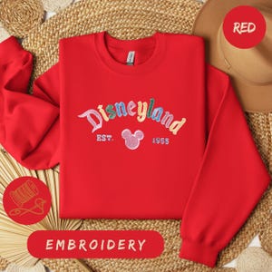 A red Disneyland sweatshirt with the text "Disneyland EST. 1955" embroidered on it, along with a red hat and straw hat. The sweatshirt is laid out on a woven mat, and there is a red embroidery label in the bottom right corner.