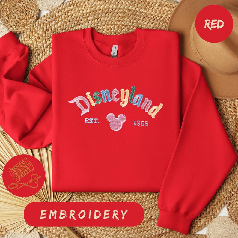 A red Disneyland sweatshirt with the text "Disneyland EST. 1955" embroidered on it, along with a red hat and straw hat. The sweatshirt is laid out on a woven mat, and there is a red embroidery label in the bottom right corner.