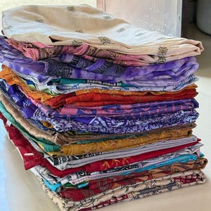Könnte beinhalten: Ein Stapel gefalteter Stoffe in verschiedenen Farben und Mustern. Die Textilien weisen Creme-, Rosa-, Lila-, Orange- und Rottöne auf. Die Stoffe sind ordentlich gestapelt, möglicherweise Schals oder ähnliche Artikel, die eine Reihe von Designs und Texturen zeigen.