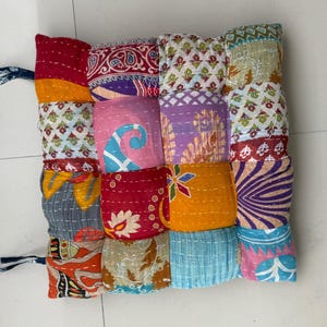 May include: A patchwork cushion with a variety of colorful fabric squares. The squares feature floral and geometric patterns in shades of red, orange, pink, blue, and purple. The cushion has a blue tie on the side.